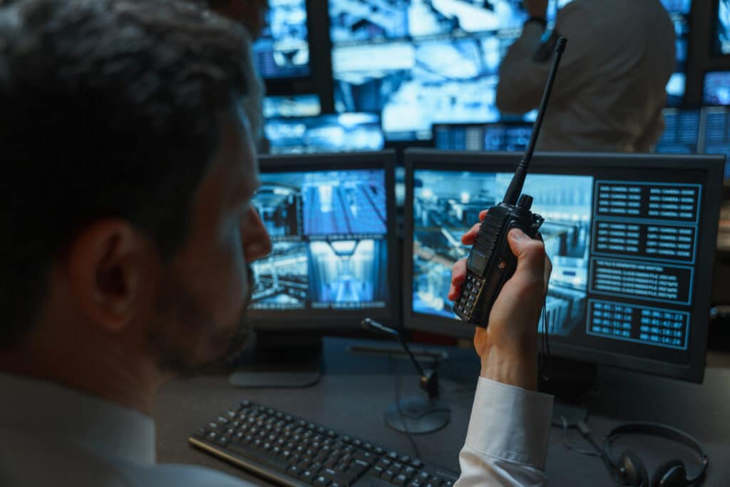 A security officer talks on a walkie talkie he is alone in the security room in complete darkness he is illuminated by monitors that display a picture from surveillance cameras